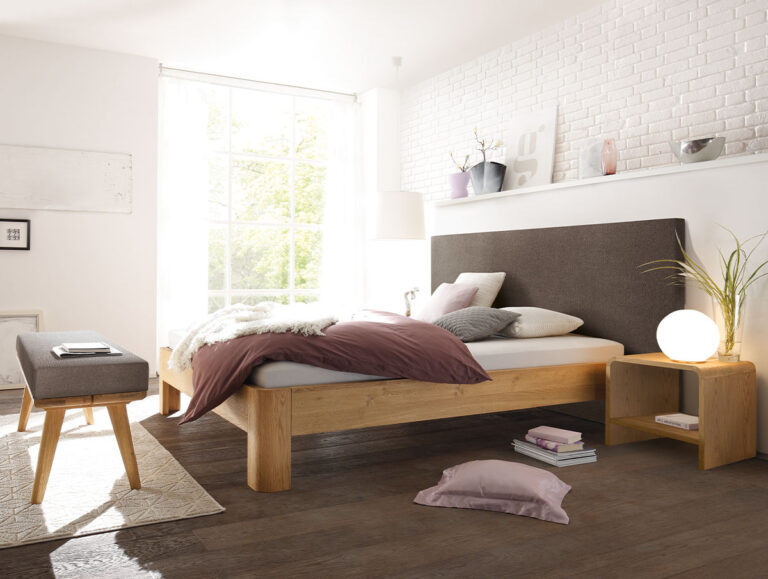 Ein modernes Schlafzimmer mit einem Holzbett, einem grauen Kopfteil, mauvefarbener Bettwäsche, einer Bank, einem Nachttisch mit einer runden Lampe und einer weißen Ziegelwand. Sonnenlicht strömt durch ein großes Fenster herein. Auf dem dunklen Holzboden liegt ein mauvefarbenes Kissen.