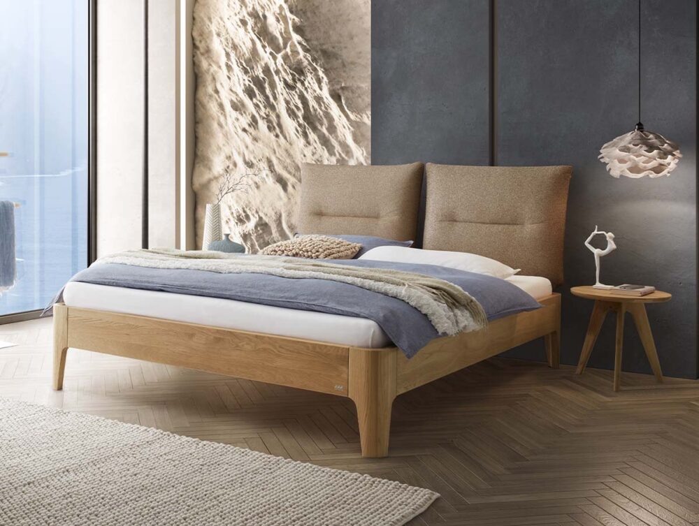 Modernes Schlafzimmer mit einem Bettgestell aus Holz, neutraler Bettwäsche, strukturierten Kissen, einer gemütlichen Decke, einem runden Nachttisch aus Holz, einer dekorativen Lampe sowie einem großen Fenster, das Tageslicht hereinlässt, und einer Akzentwand aus Stein.