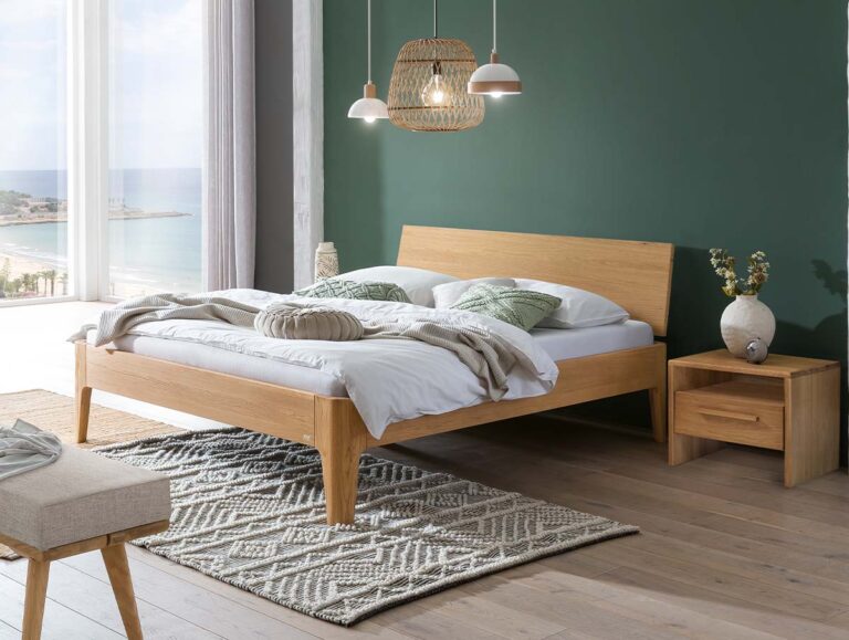 Modernes Schlafzimmer mit einem Holzbett, einer grünen Akzentwand, Hängelampen, einem Nachttisch mit Vase und einem großen Fenster mit Blick auf die Küste. Ein strukturierter Teppich und gemütliche Bettwäsche runden die ruhige, minimalistische Einrichtung ab.