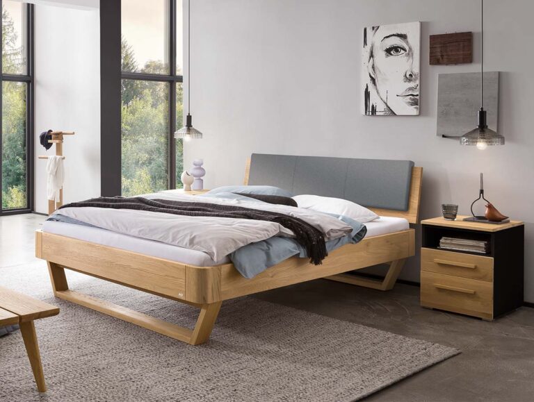 Ein modernes Schlafzimmer mit dem Hasena-Massivholzbett „Eiche-Bianco“ 9.106 mit Holzrahmen, grauem Kopfteil, ordentlich gemachtem Bett, einem Nachttisch mit Schubladen, hängenden Pendelleuchten und zeitgenössischer Wandkunst. Große Fenster lassen Tageslicht herein.