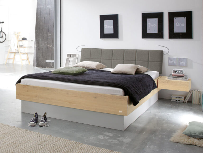 Modernes Schlafzimmer mit einem Holzbett, einem grauen Kopfteil, einer schwarzen Decke, Kissen, einem schwebenden Nachttisch, Turnschuhen auf dem Boden und drei gerahmten Drucken an einer weißen Wand.