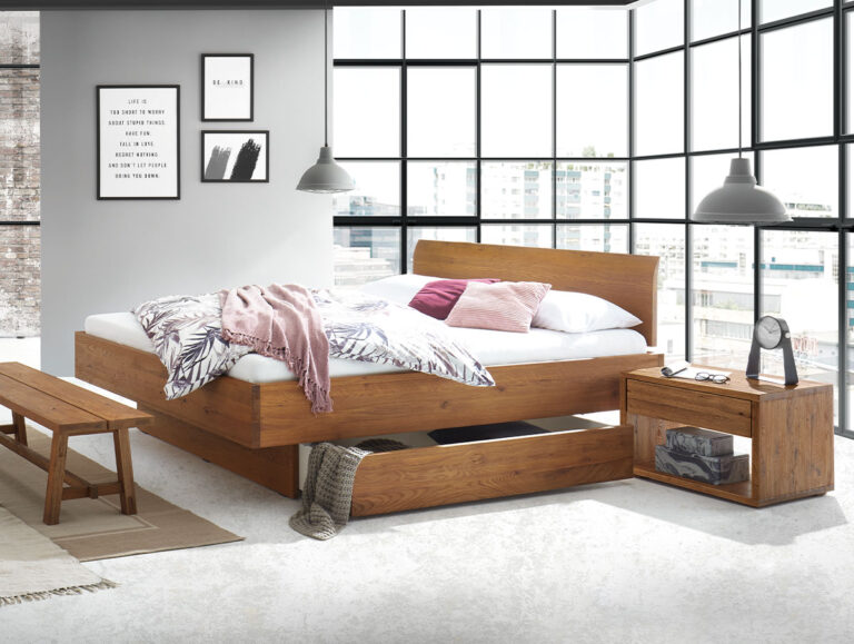 Modernes Schlafzimmer mit großen Fenstern, einem Holzbett, einem Nachttisch und einer Bank. Rosa und tropisch anmutende Bettwäsche, Wandbilder und zwei Pendelleuchten. Auf dem Nachttisch stehen eine Uhr, Bücher und eine Glasvitrine. Blick auf die Stadtlandschaft.