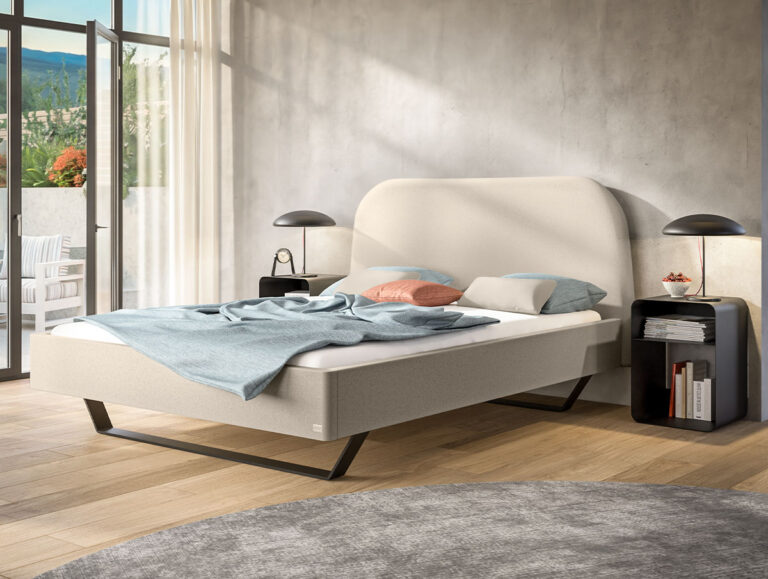 Modernes Schlafzimmer mit einem beigen Polsterbett, einer blauen Decke und Kissen. Auf den beiden schwarzen Nachttischen stehen passende Lampen. Große Fenster sorgen für Tageslicht und bieten einen Blick auf eine begrünte Terrasse.