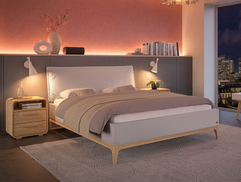 Modernes Schlafzimmer mit einem Holzbett, beiger und malvenfarbener Bettwäsche, einem Nachttisch mit Lampe und Deko sowie Blick auf die Stadt durch das Fenster. Eine warme Raumbeleuchtung setzt die rosa Akzentwand in Szene.