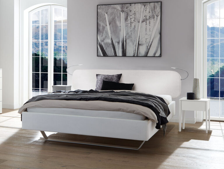 Ein modernes Schlafzimmer mit einem weißen Bett, Kissen, grauer Bettwäsche, einem Nachttisch mit Deko, großen Fenstern und einem monochromen Gemälde mit Naturmotiv an der Wand. Sonnenlicht strömt herein und schafft eine helle, luftige Atmosphäre.