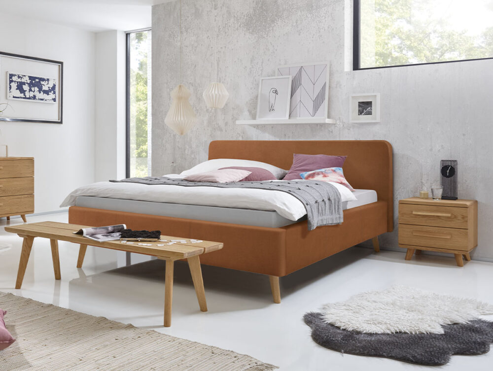 Modernes Schlafzimmer mit einem braunen Polsterbett, einer Holzbank, einem Nachttisch und einer Kommode. Neutrale Einrichtung, strukturierter Teppich, Wanddekoration, Regal und großes Fenster, durch das Tageslicht hereinströmt. Hellgraue Wände und minimalistisches Design.
