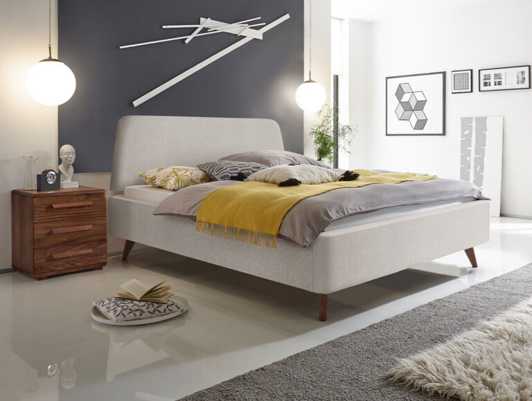 Modernes Schlafzimmer mit einem beigen Bett, einer gelben Decke, zwei Pendelleuchten, einem Nachttisch aus Holz, geometrischen Wandbildern und Dekoartikeln. Der Raum ist minimalistisch eingerichtet und in neutralen Farbtönen gehalten.