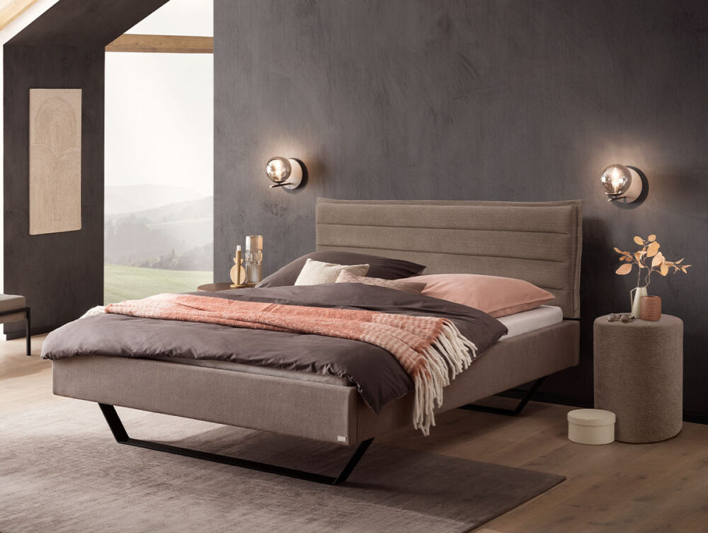 Modernes Schlafzimmer mit einem grauen Polsterbett, rosa-brauner Bettwäsche, Wandleuchten, einem runden Beistelltisch mit einer Vase und einem großen Fenster, das einen malerischen Ausblick bietet. Die Wände und der Boden sind in neutralen Farbtönen gehalten.