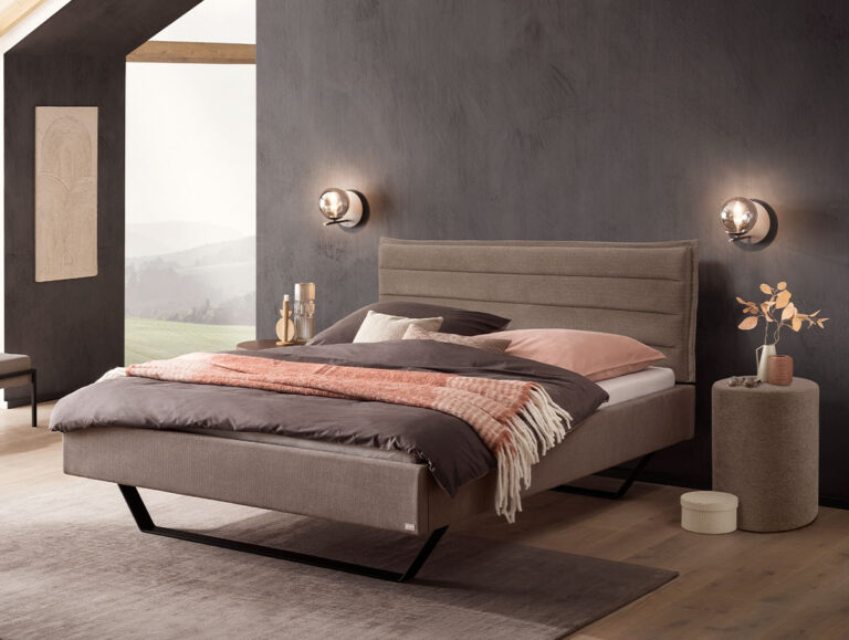 Modernes Schlafzimmer mit einem grauen Polsterbett, rosa-brauner Bettwäsche, Wandleuchten, einem runden Beistelltisch mit einer Vase und einem großen Fenster, das einen malerischen Ausblick bietet. Die Wände und der Boden sind in neutralen Farbtönen gehalten.