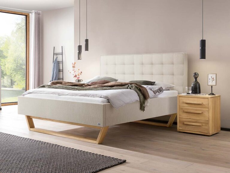 Ein modernes Schlafzimmer mit einem beigen Polsterbett, einem Holzrahmen, ordentlich gemachter Bettwäsche, einem hölzernen Nachttisch mit Deko und einem großen Fenster, durch das Tageslicht hereinströmt.