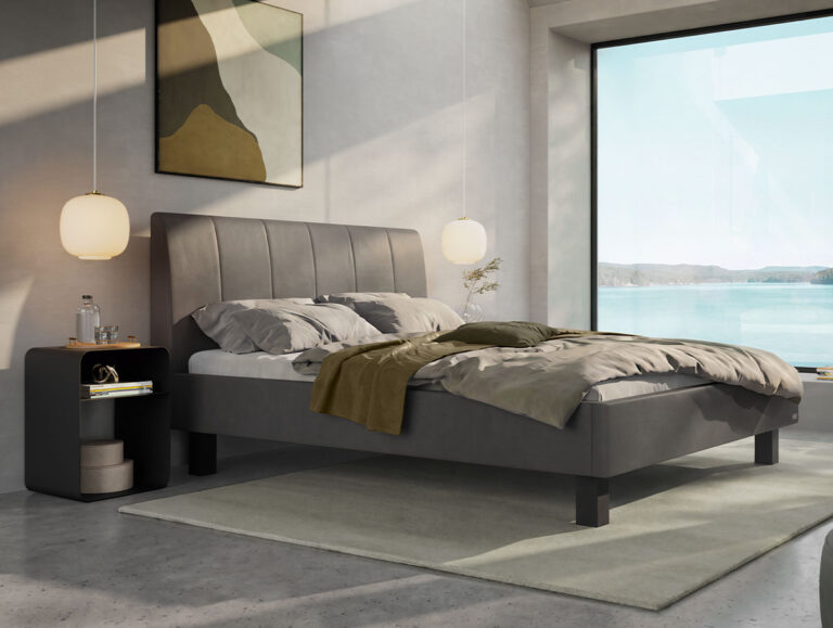 Ein modernes Schlafzimmer mit einem großen grauen Bett, unordentlicher Bettwäsche, einem Nachttisch mit einem Glas Wasser, Hängeleuchten, abstrakten Wandbildern und einem großen Fenster, das den Blick auf das Wasser und ferne Hügel freigibt.