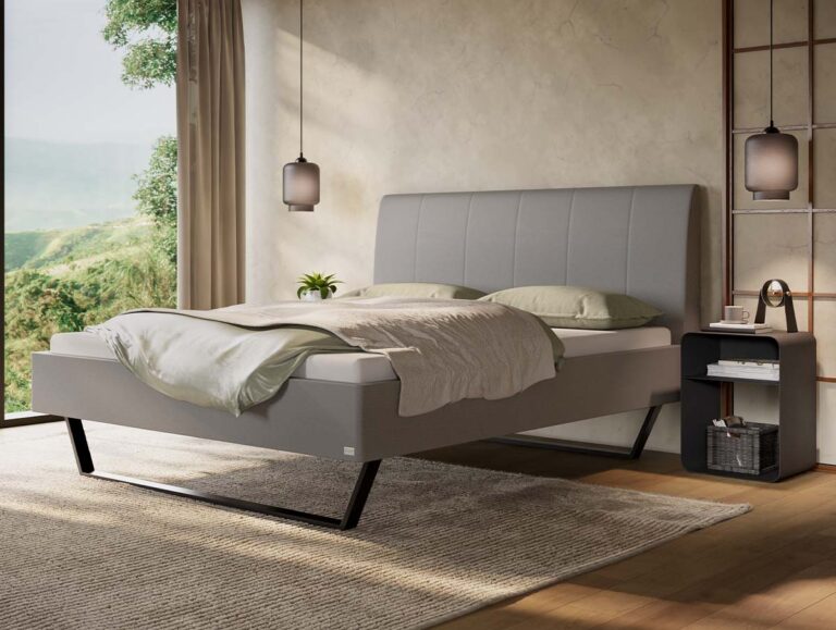 Modernes Schlafzimmer mit einem grauen Polsterbett, heller Bettwäsche, einem Nachttisch mit Lampe und Wecker, einem großen Fenster mit Blick ins Grüne sowie einer Einrichtung in neutralen Farbtönen.