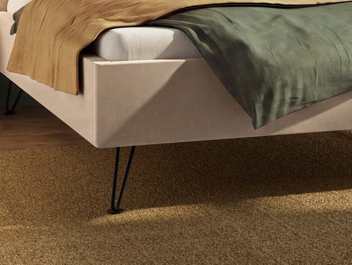 Eine Nahaufnahme eines modernen beigen Bettgestells mit schwarzen Metallbeinen, das mit einer grünen Decke und einem braunen Laken bedeckt ist und auf einem strukturierten beigen Teppich steht.