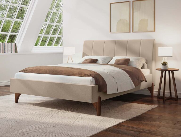 Modernes Schlafzimmer mit einem beigen Polsterbett, weißer und brauner Bettwäsche, zwei Nachttischen mit Lampen, einem großen Teppich, Holzboden und schrägen Fenstern, durch die Tageslicht hereinfällt.
