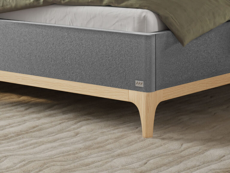 Auf einem strukturierten, sandfarbenen Teppich steht ein modernes Bett mit hellen Holzbeinen und einem grau gepolsterten Rahmen. Über eine Ecke des ordentlich gemachten Bettes ist eine grünliche Decke drapiert.