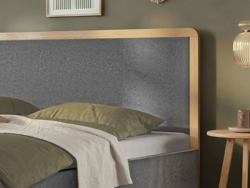 Ein ordentlich gemachtes Bett mit grünen Kissen und einer grünen Decke, ein graues, gepolstertes Kopfteil mit Holzrahmen, ein kleiner runder Nachttisch, eine hängende Glaslampe und ein gerahmtes Bild an einer grünen Wand.
