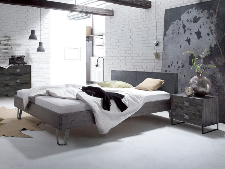 Hasena Industrial-Massivholzbett Factory-Line 5.34 – ein flaches Plattformbett mit dunklem Holzrahmen, grauem gepolstertem Kopfteil und Metallbeinen im Haarnadelstil, dazu ein passender Nachttisch in einem Schlafzimmer im Loft-Stil.