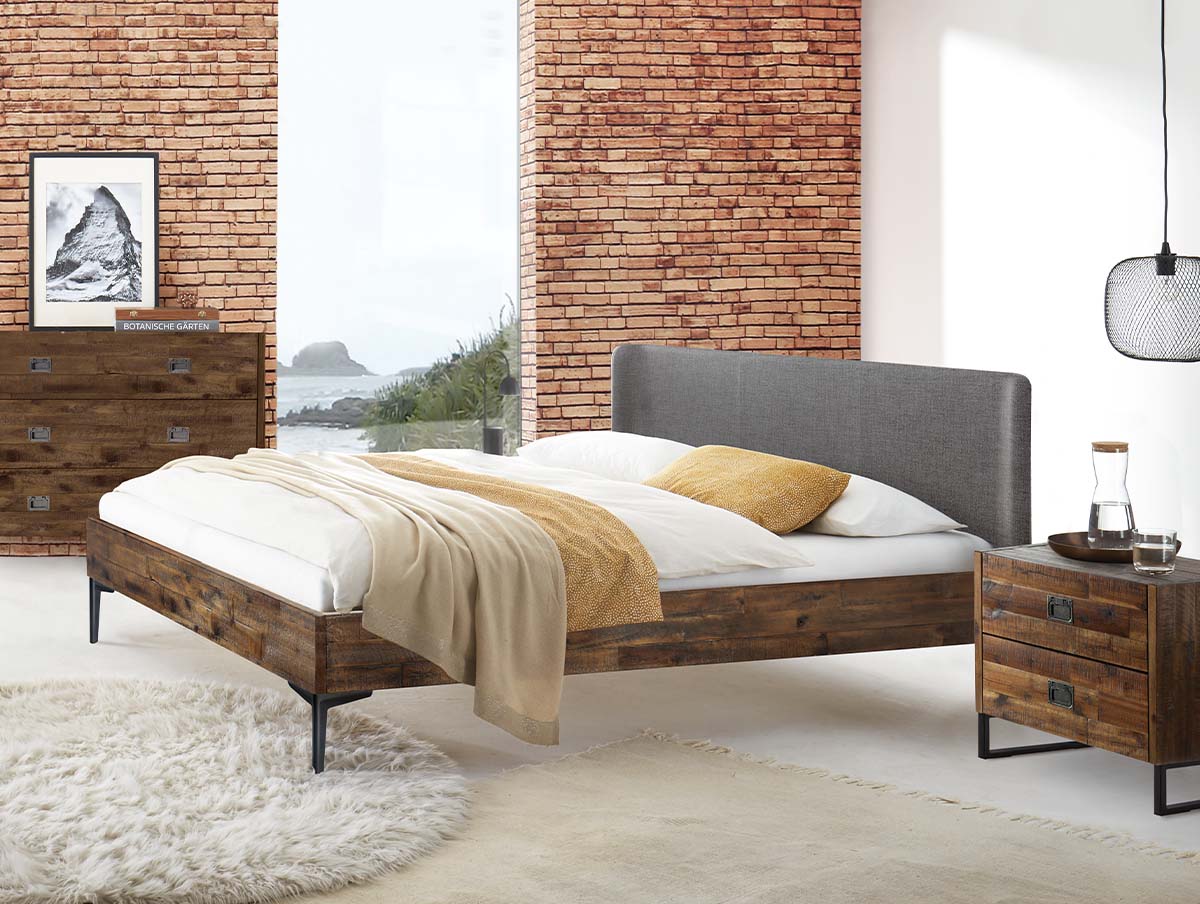 Ein Bett mit Holzrahmen und grauem, gepolstertem Kopfteil in einem Schlafzimmer mit einer Backsteinwand und passenden Holzmöbeln.