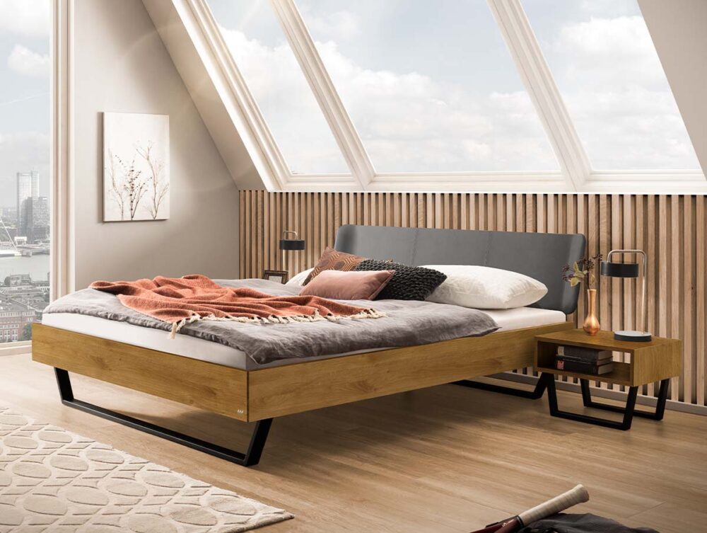 Ein Bettgestell aus Holz mit einem grauen, gepolsterten Kopfteil und Metallbeinen, das in einem Raum mit einem großen, schräg verlaufenden Oberlicht und einer Wandverkleidung aus Holzlatten steht.