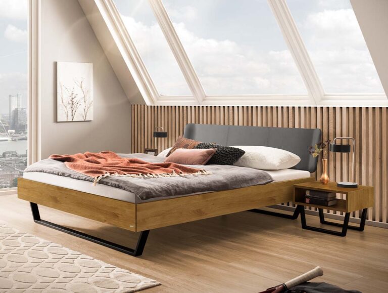 Ein Bettgestell aus Holz mit einem grauen, gepolsterten Kopfteil und Metallbeinen, das in einem Raum mit einem großen, schräg verlaufenden Oberlicht und einer Wandverkleidung aus Holzlatten steht.