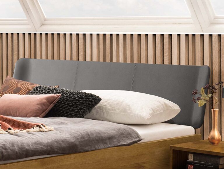 Ein Bett mit einem grauen, gepolsterten Kopfteil, einer Wandverkleidung aus Holzlatten und Kissen in Weiß-, Schwarz- und Rosatönen.