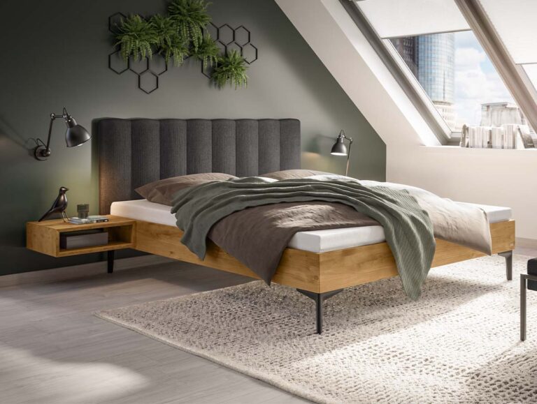 Ein Plattformbett mit einem dunkelgrauen, gepolsterten Kopfteil und einem hellen Holzrahmen, ergänzt durch einen schwebenden Nachttisch und mehrlagige Bettwäsche in einem Schlafzimmer im Dachgeschoss.