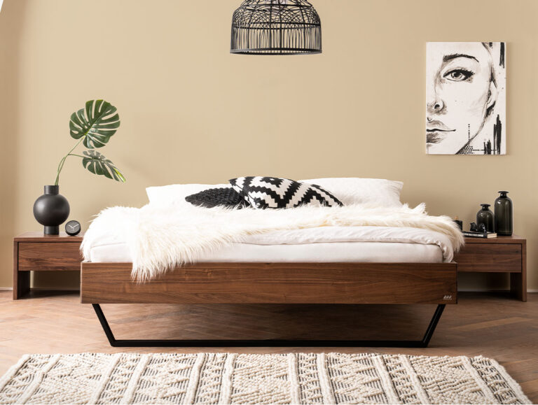 Ein Bettgestell aus Holz mit schwarzen Metallbeinen, bezogen mit weißer Bettwäsche, Kissen und einer Decke aus Kunstfell.
