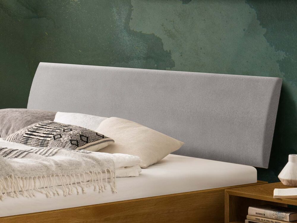 Ein hellgraues, gepolstertes Kopfteil hinter einem Bett mit mehreren Kissen und einer Zudecke.