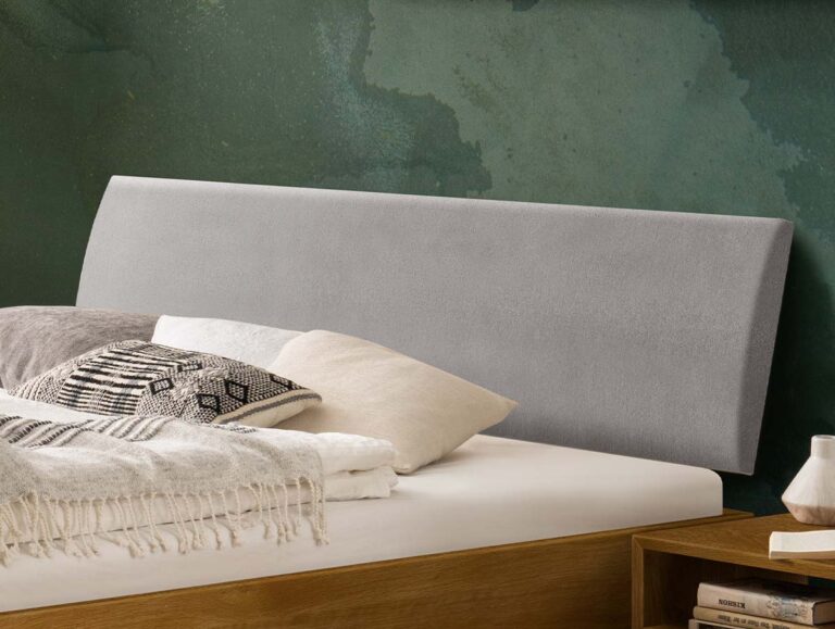 Ein hellgraues, gepolstertes Kopfteil hinter einem Bett mit mehreren Kissen und einer Zudecke.