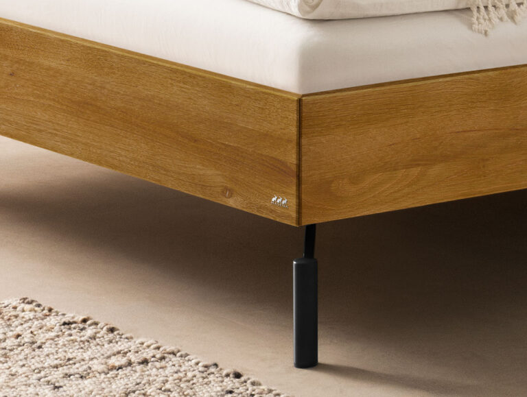 Ein Bettgestell aus Holz mit einer weißen Matratze und einem einzelnen schwarzen, zylindrischen Bein.