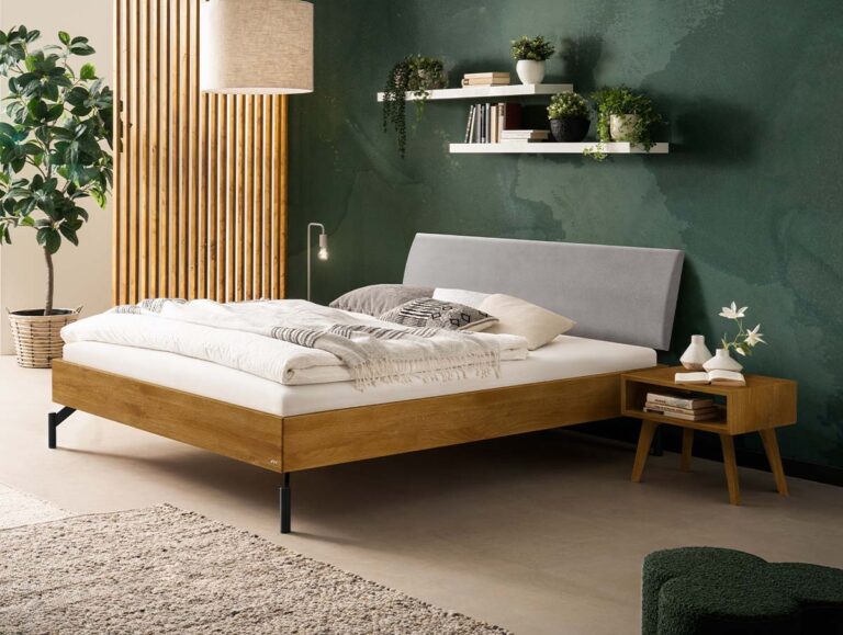 Ein Bettgestell aus Holz mit einem grauen, gepolsterten Kopfteil in einem Schlafzimmer mit dunkelgrüner Wand, schwebenden Regalen und einem Nachttisch aus Holz.