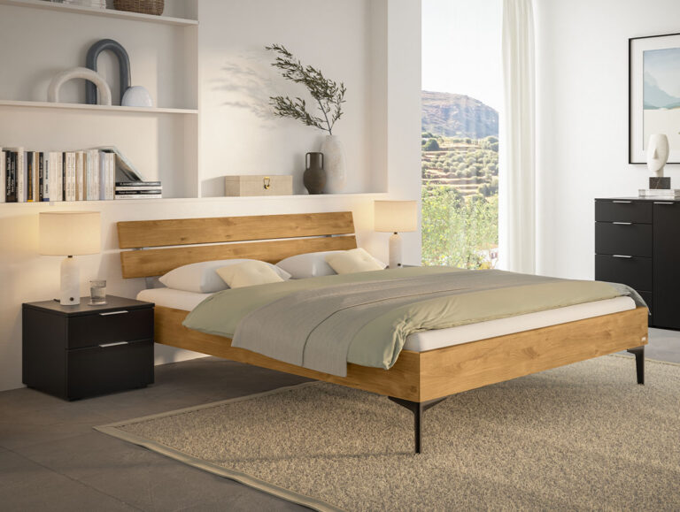 Ein Bettgestell aus hellem Holz mit einer grünen Bettdecke, flankiert von zwei schwarzen Nachttischen in einem hellen Schlafzimmer.