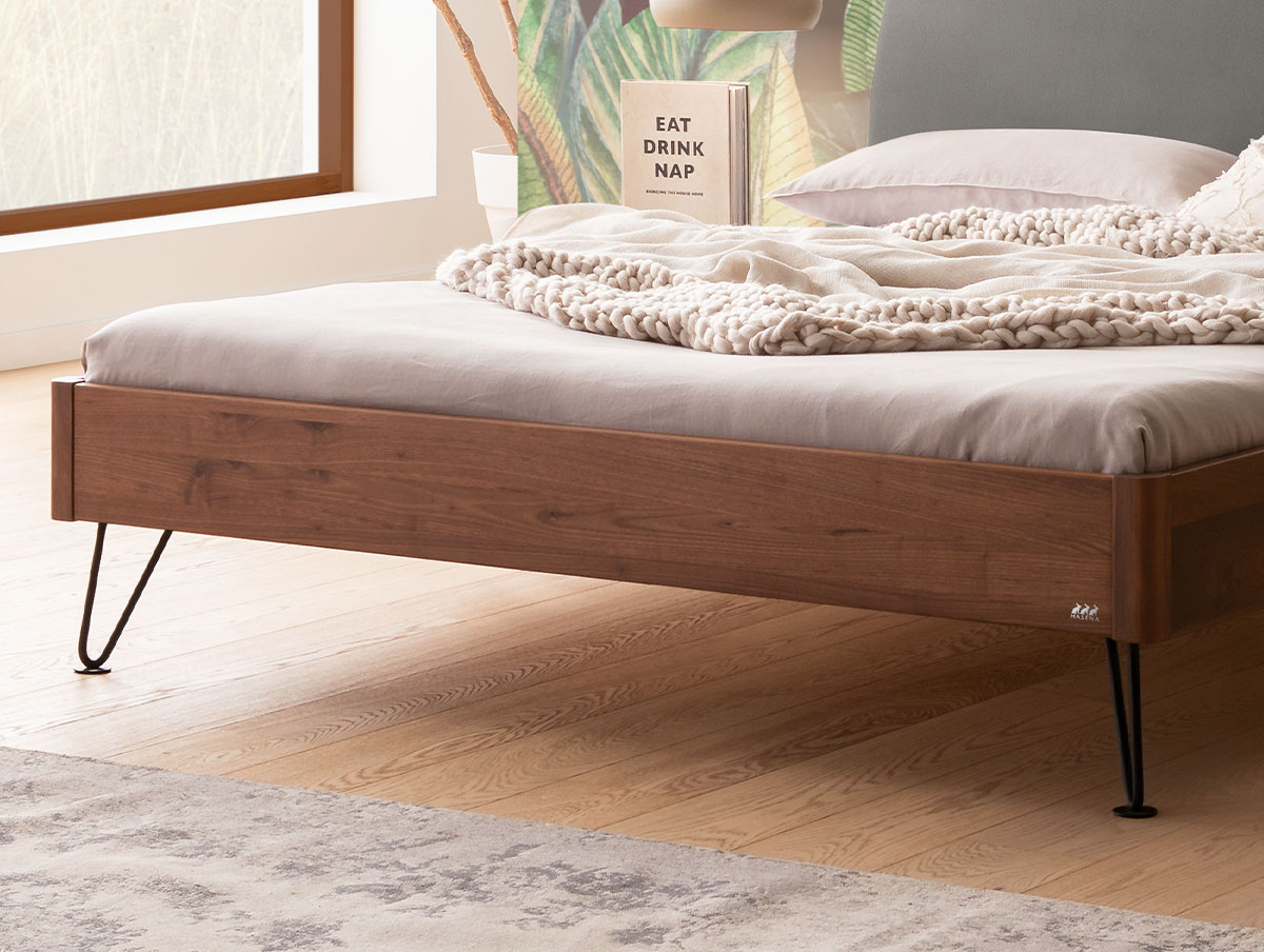 Ein Plattformbett mit Holzrahmen und schwarzen Metallbeinen in Haarnadelform, bedeckt mit einer dicken Strickdecke und einem Buch mit dem Titel Eat Drink Nap.